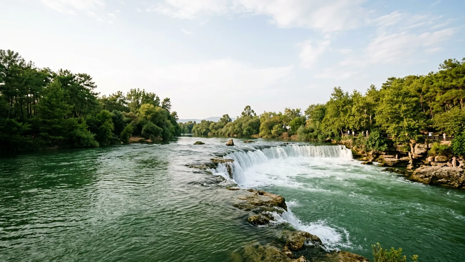 Manavgat, Türkiye — the Manavgat river, a wide low waterfall spilling over a broad rock ledge into green water with pine woods along both banks.