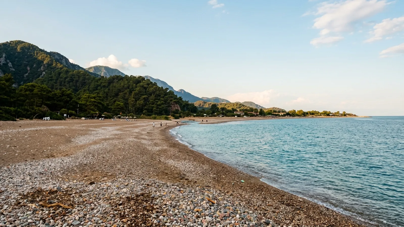 Cirali, Türkiye — the wide pebble beach at Cirali below wooded mountains, a broad open bay with a stream mouth and unspoilt low-rise surroundings.